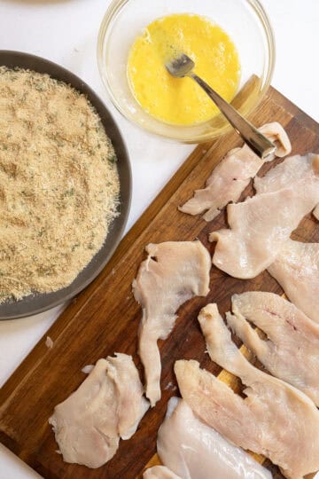 Thinly sliced chicken breast laid out on cutting board with bowl of whisked eggs and seasoned breadcrumbs.