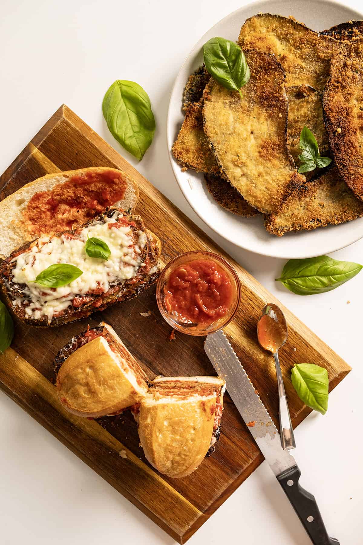 Two eggplant parm sandwiches on a cutting board with a plate of fried eggplant.