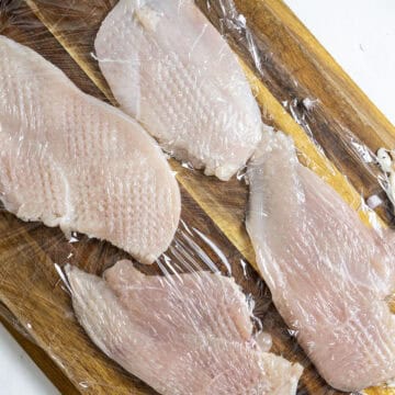 Sliced chicken breast with saran wrap over it being tenderized.