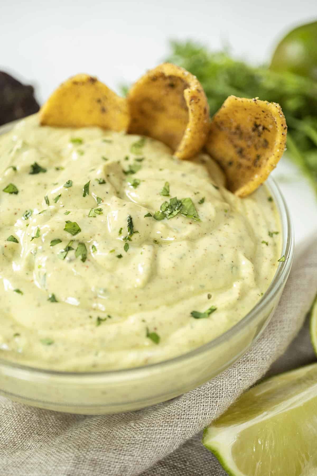 avocado lime crema in a glass bowl with limes and cilantro on side and chips in the crema.
