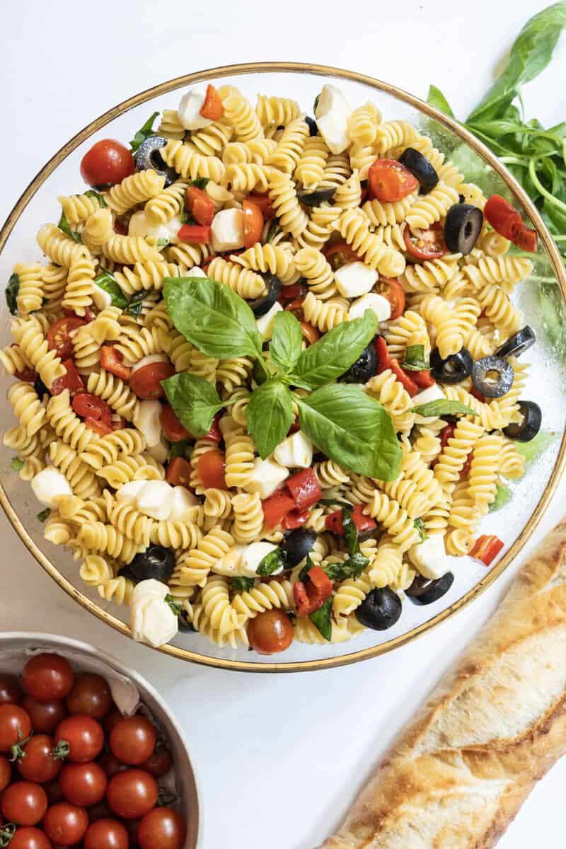 Italian pasta salad in a large bowl with garnish of basil, on the side is a loaf of bread and bowl of cherry tomatoes.