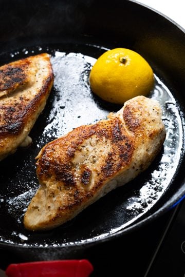 Golden brown chicken breast in cast iron skillet with lemon.