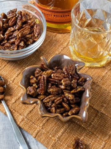 Candied pecans in 2 separate bowls next to a glass of bourbon.