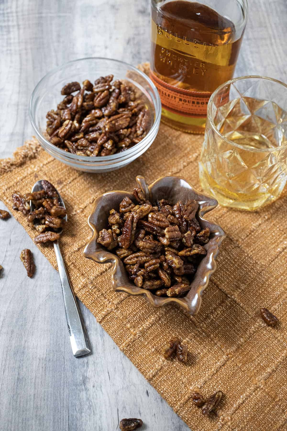 Candied pecans in 2 separate bowls next to a glass of bourbon.