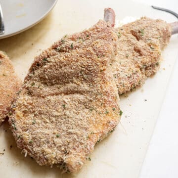 Pork chops dredged in bread crumbs on a cutting board.