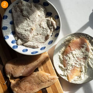 Dredging chicken breast in flour on a plate.