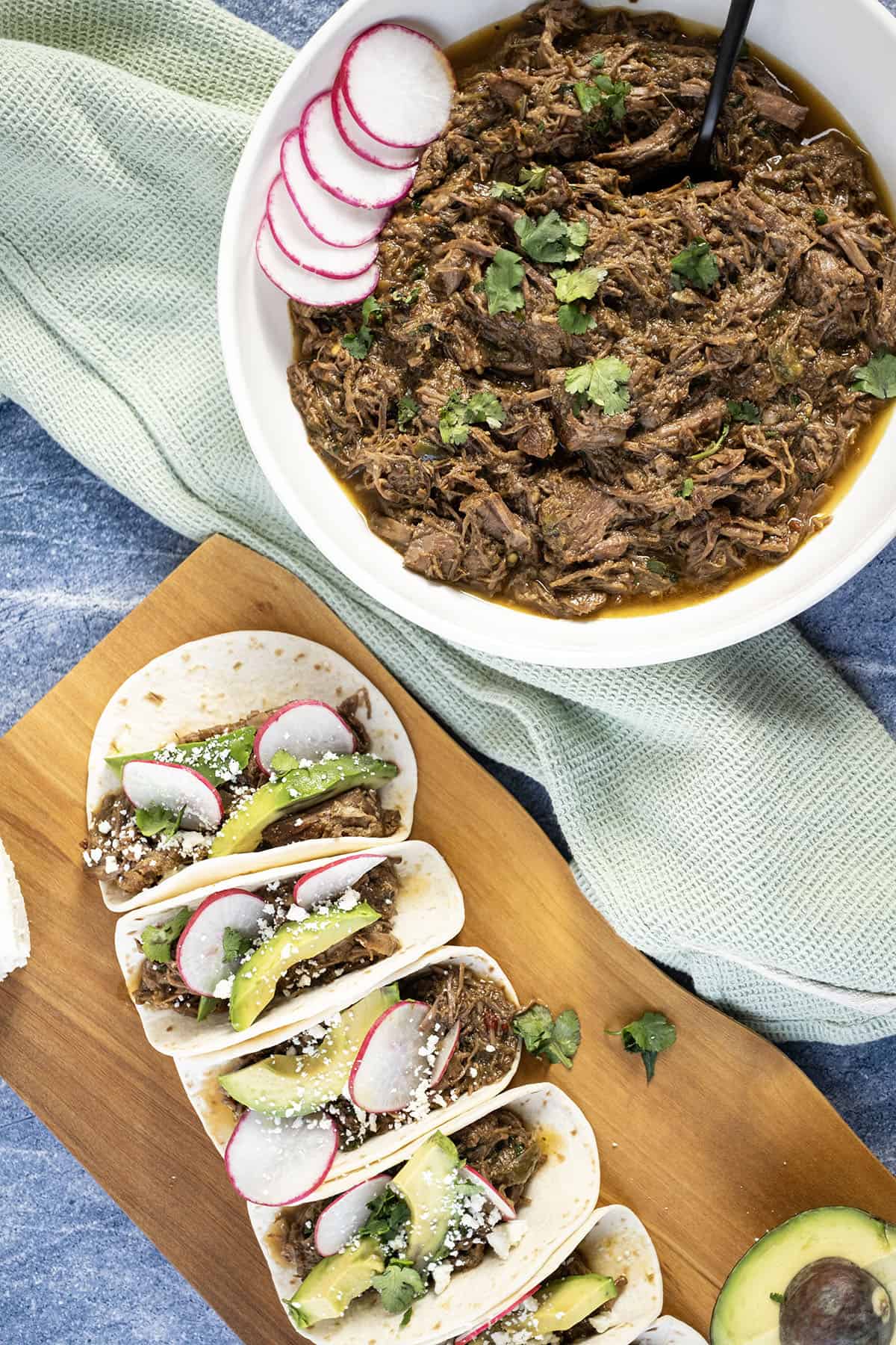 Beef barbacoa in a bowl alongside tacos with sliced radish, avocado, and cotija cheese.
