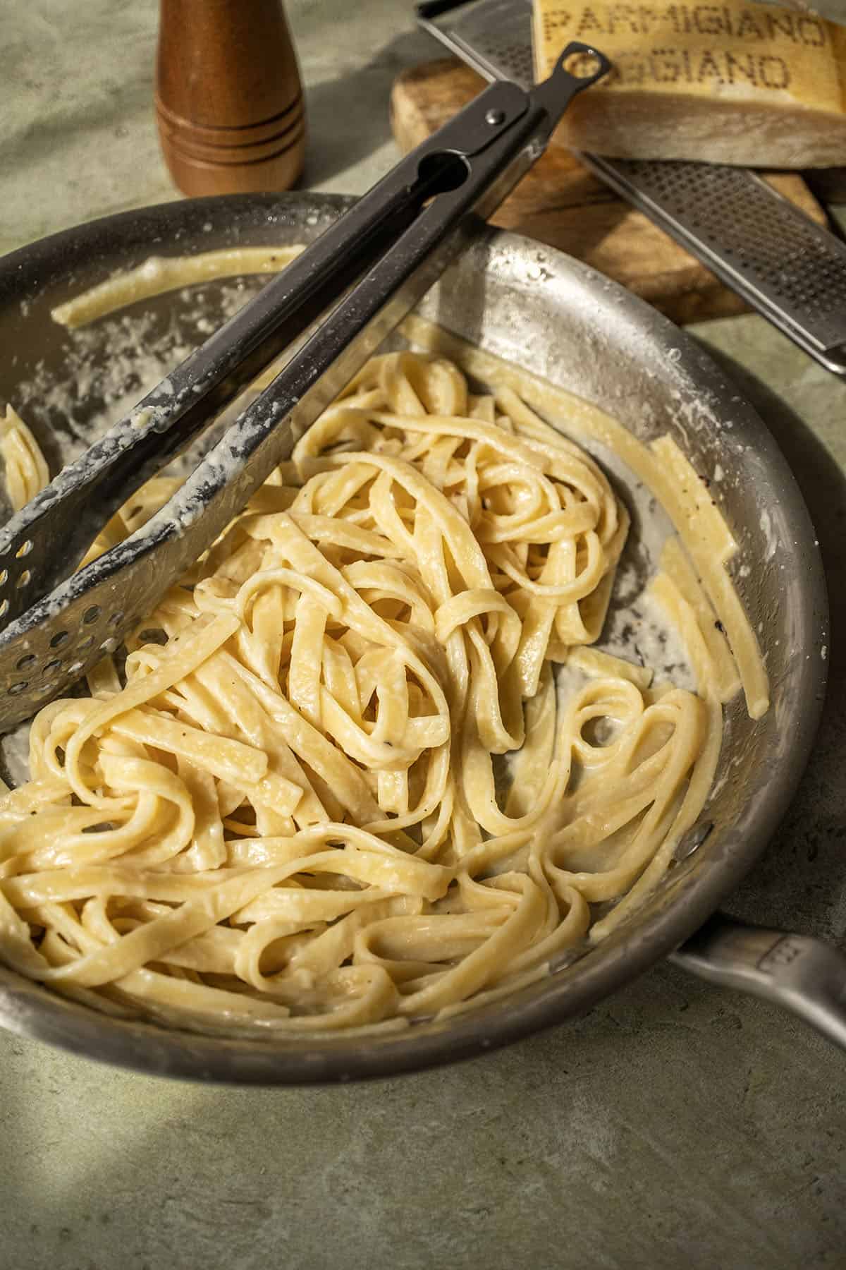 Fettucine in alfredo sauce in a pan with tongs.