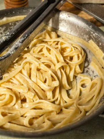 Fettucine in alfredo sauce in a pan with tongs.
