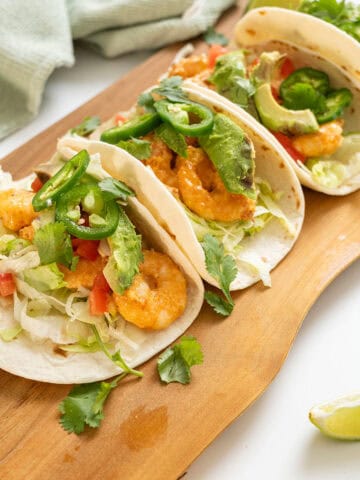 Volcano shrimp tacos on a cutting board topped with cilantro, tomatoes, jalapeno, and avocado.