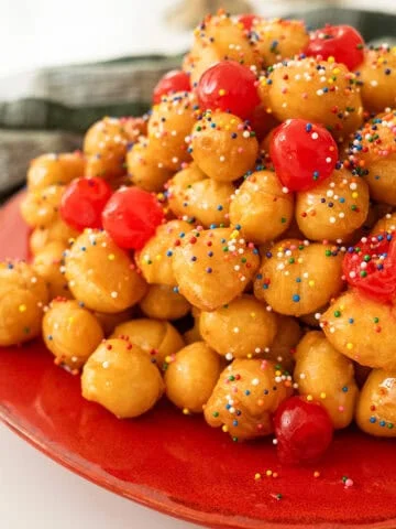 Struffoli piled into a pyramid topped with non-pareils and maraschino cherries.