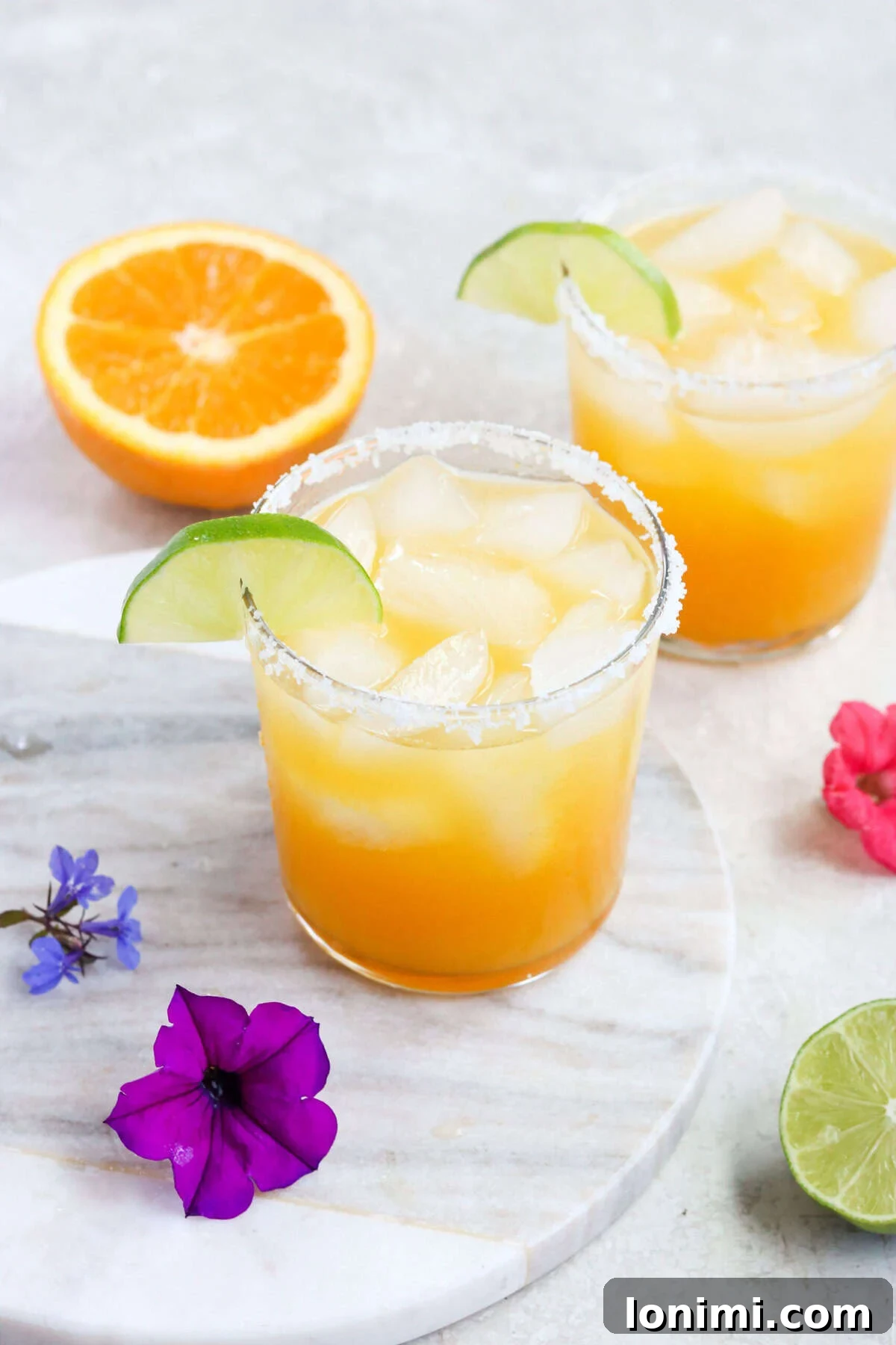 Two clear glasses filled with vibrant yellow passion fruit margaritas, garnished with lime wedges and purple flowers, sitting on a table next to a sliced orange.