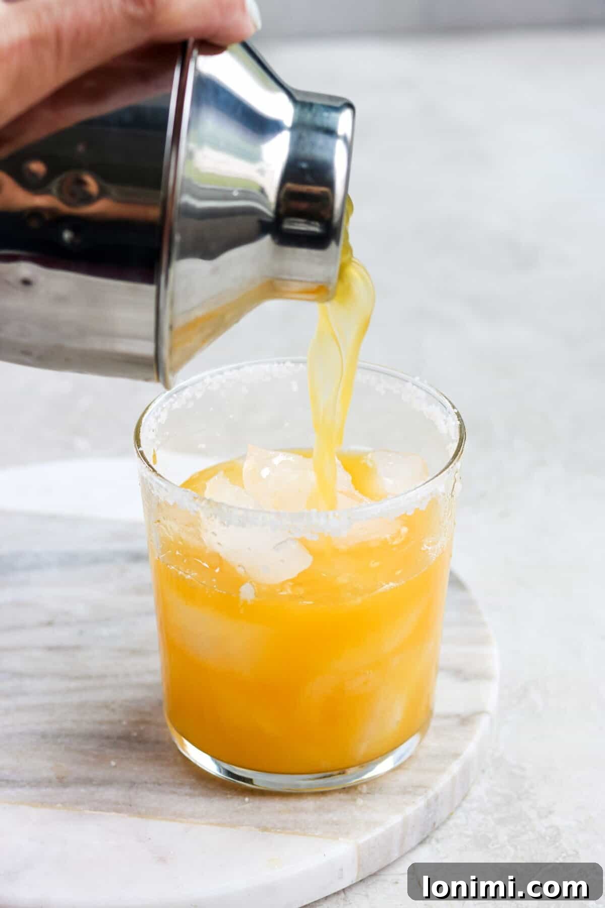 A cocktail shaker expertly pouring a passion fruit margarita into a glass filled with ice and a perfectly salted rim.