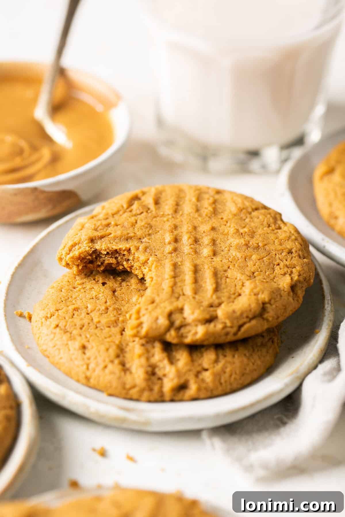Mary's Signature Gluten-Free Peanut Butter Cookies 7 Two perfect gluten-free peanut butter cookies stacked on a white plate, with a background of a bowl filled with creamy peanut butter, emphasizing their deliciousness.