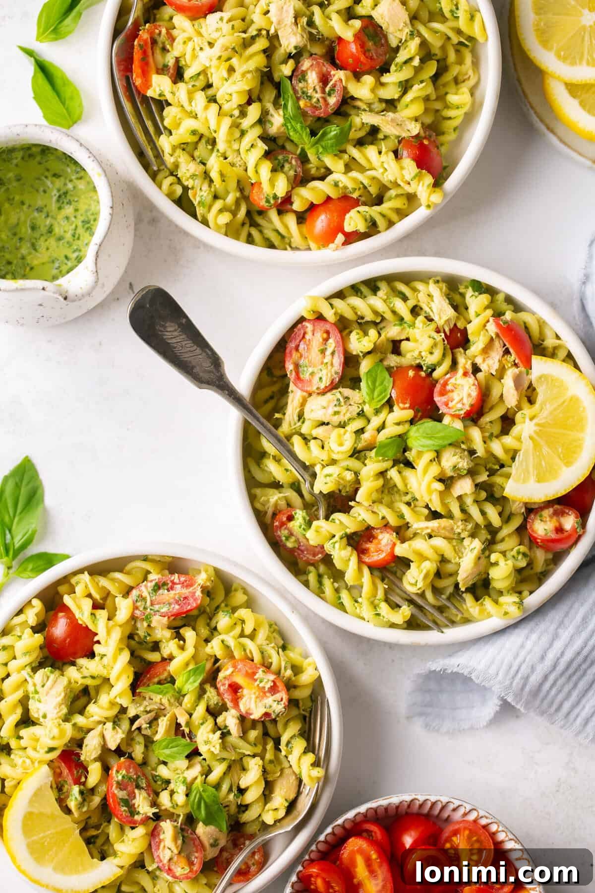 three white bowls of pesto tuna pasta with tomatoes next to a saucer with homemade pesto sauce