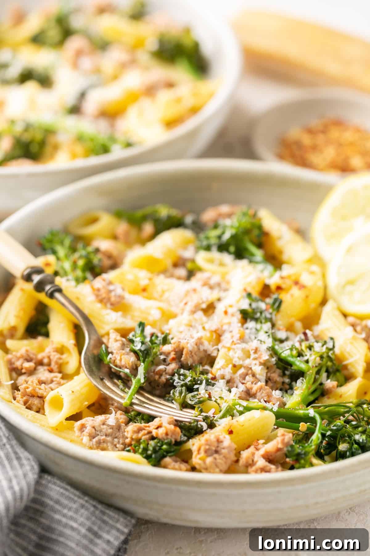 A white bowl filled with sausage broccolini pasta, garnished with a fresh lemon wedge.
