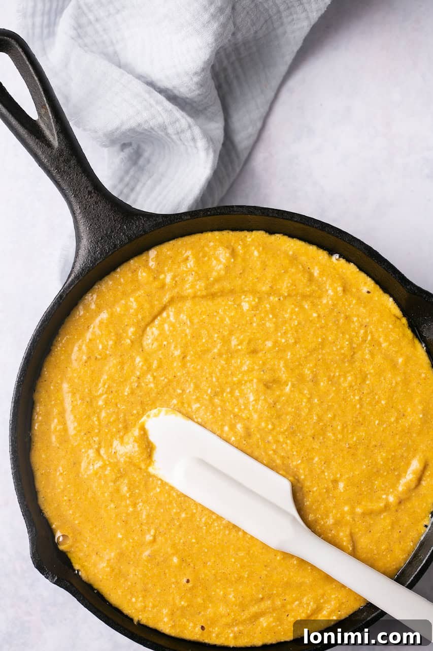 Golden Sweet Potato Cornbread 5 Raw sweet potato cornbread batter resting in a cast iron pan, with a white spatula lying on top, ready to go into the oven for baking.