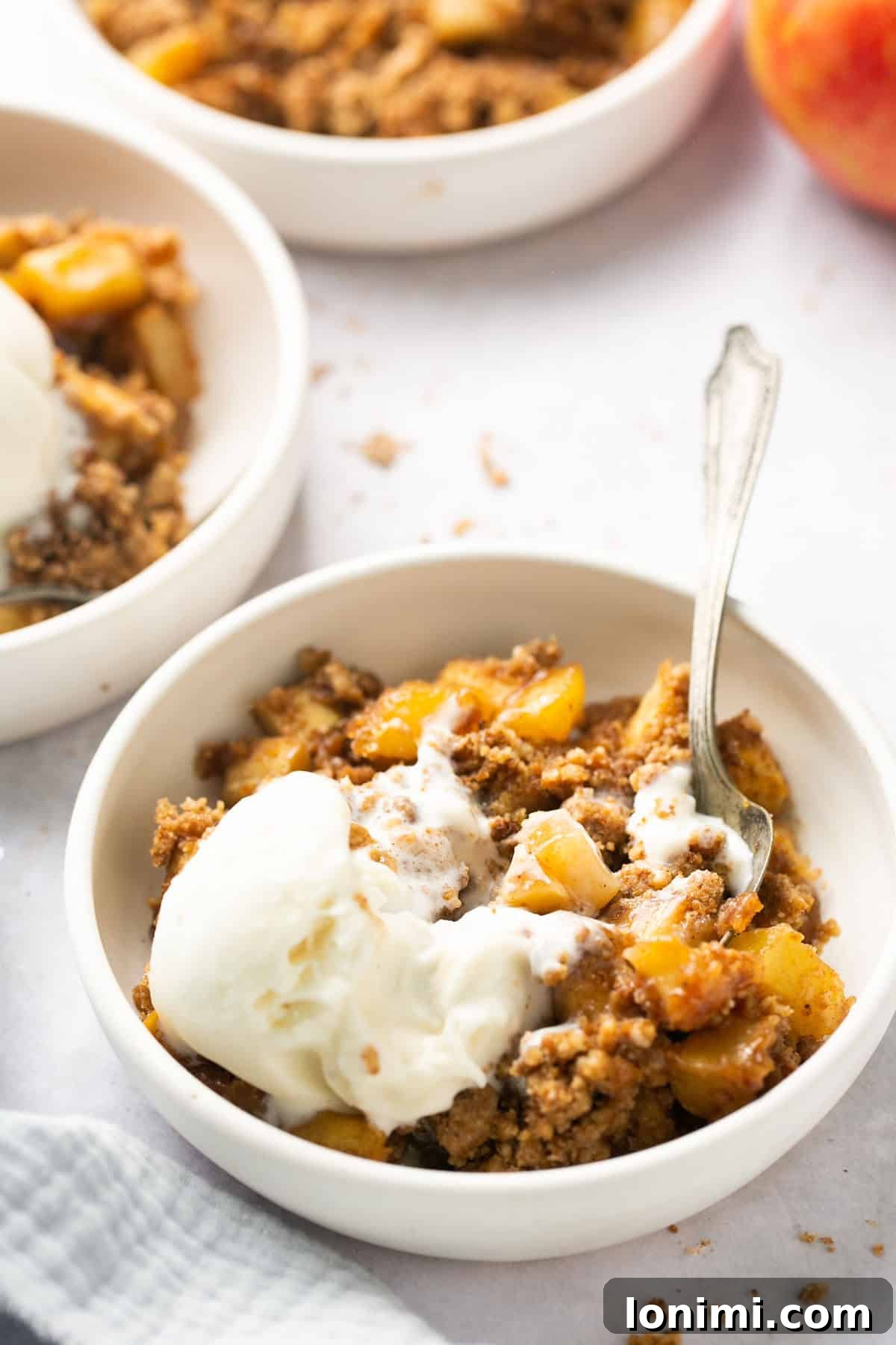 a slice of healthy apple crumble in a white bowl topped with ice cream and a silver spoon, ready to be enjoyed