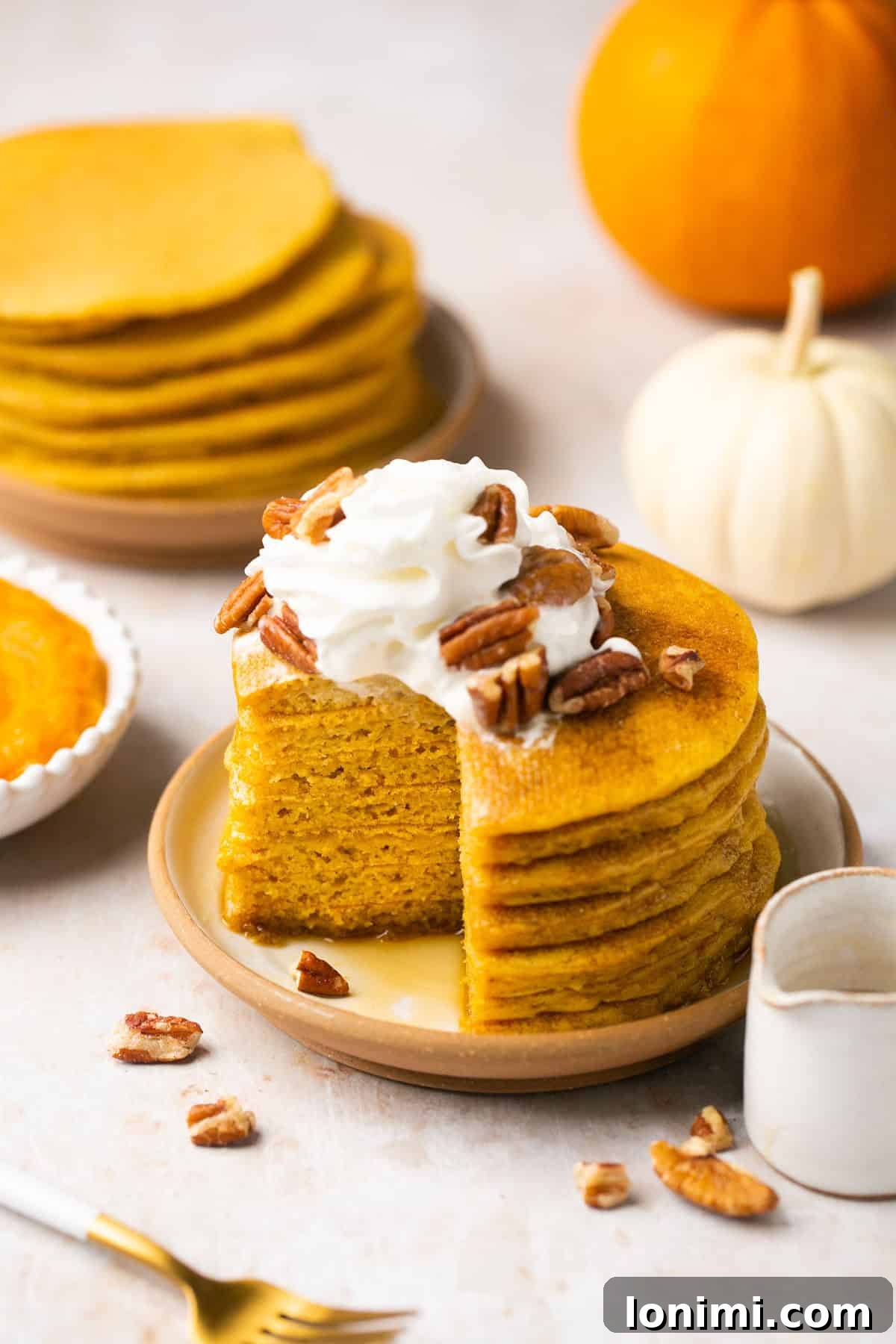 gluten free pancakes stacked on a plate with whipped cream and pecans with pumpkins and more pancakes in the background