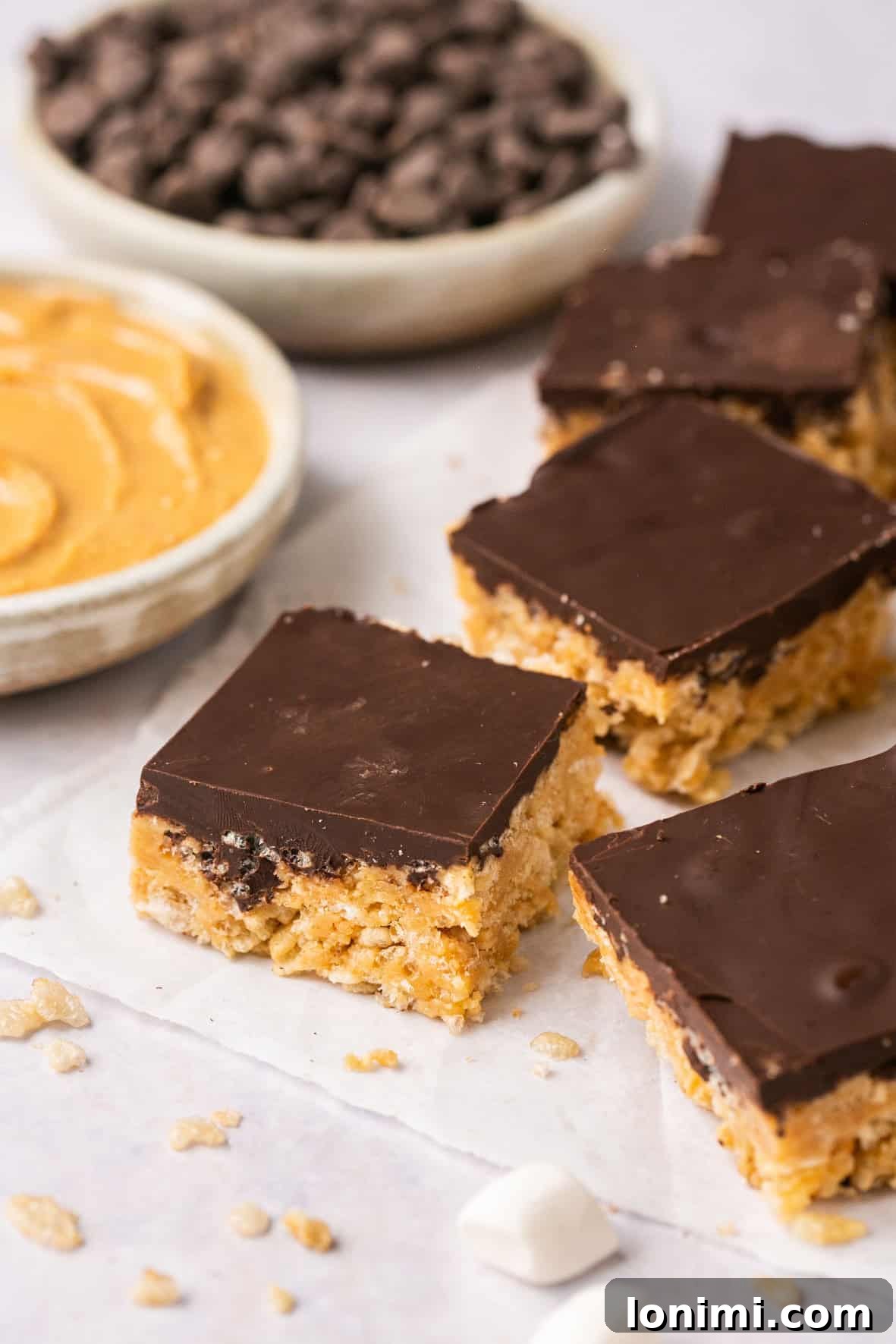 chocolate peanut butter rice krispie treats on parchment paper with peanut butter and chocolate chips in the background