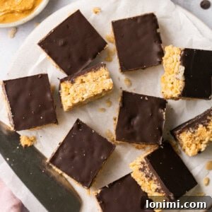 chocolate peanut butter rice krispie treats laid out on parchment paper with a bowl of peanut butter and marshmallows in the background