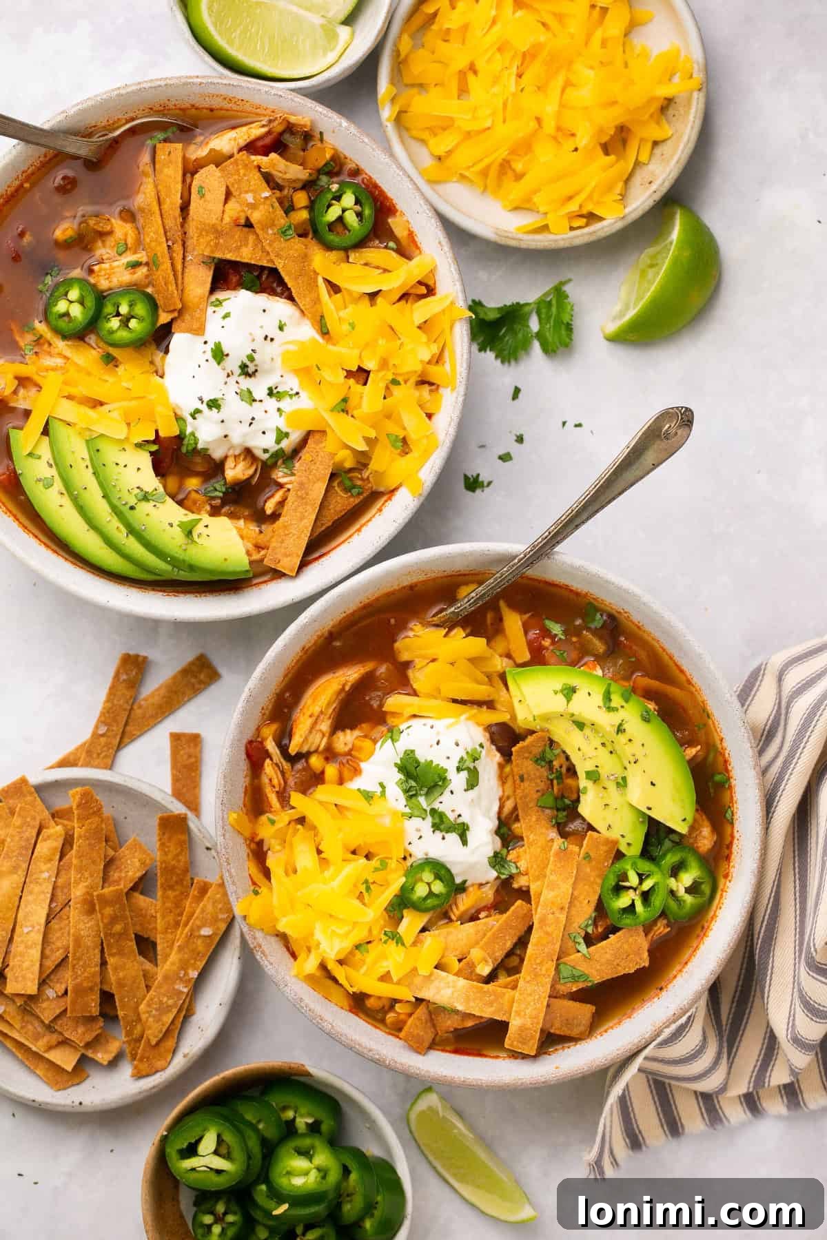 Chick-fil-A Inspired Tortilla Soup 6 An overhead shot of two white bowls filled with chicken tortilla soup and topped with homemade tortilla strips, avocado, sour cream, and shredded cheese next to small nesting bowls with shredded cheese, jalapeños, and tortilla strips