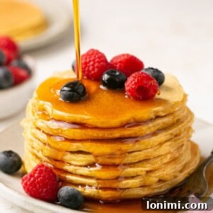 protein pancakes stacked on a plate with berries and maple syrup being poured on top