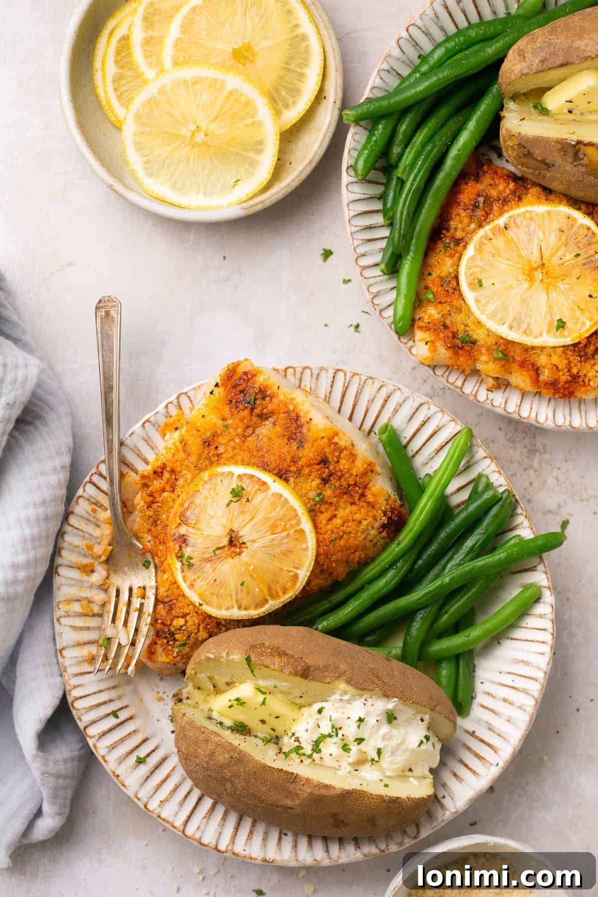 An overhead shot of two plates with air fryer halibut topped with lemon slices next to a serving of green beans and baked potato, showcasing a complete and delicious meal.