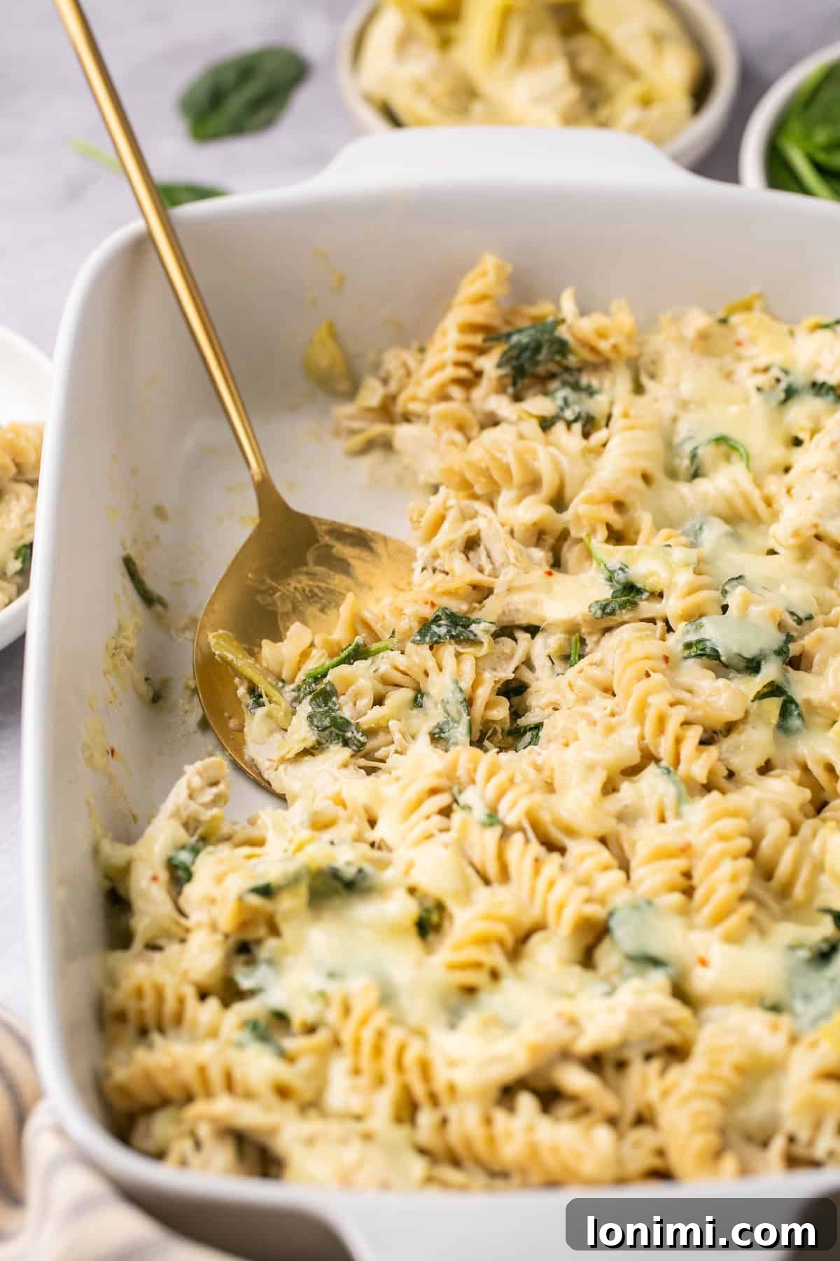 Spinach Artichoke Chicken Casserole Vegan Adaptable 7 A gold serving spoon scooping out a generous serving of creamy spinach artichoke chicken pasta from a baking dish.