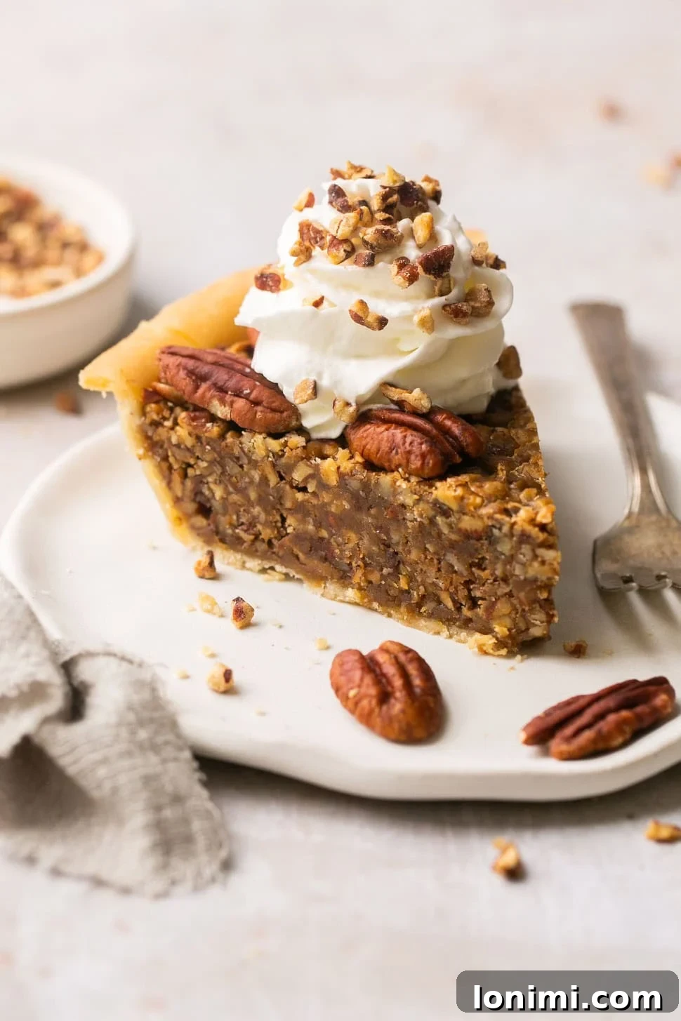 A perfectly baked slice of gluten-free pecan pie, elegantly presented on a white plate, adorned with a dollop of fluffy whipped cream and a sprinkle of chopped pecans.