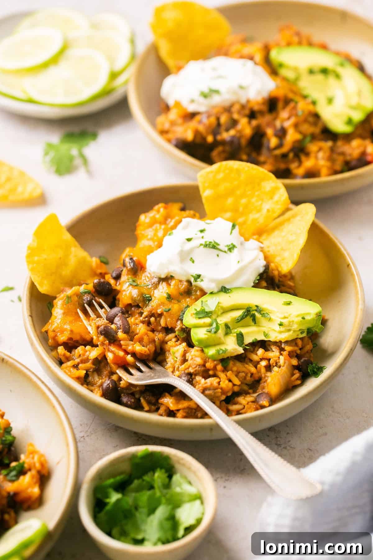 A vibrant bowl filled with hearty beef taco in a bowl recipe ingredients, lavishly garnished with creamy avocado slices, a generous dollop of sour cream, and fresh cilantro. A silver fork rests invitingly on the side of the bowl.
