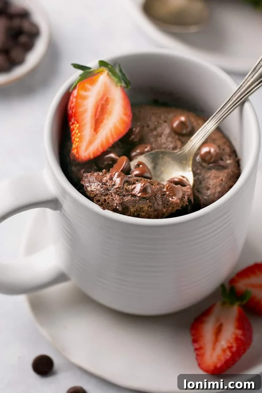 A delicious protein chocolate mug cake topped with chocolate chips and a fresh strawberry slice, with a spoonful being taken out, highlighting its fudgy texture.