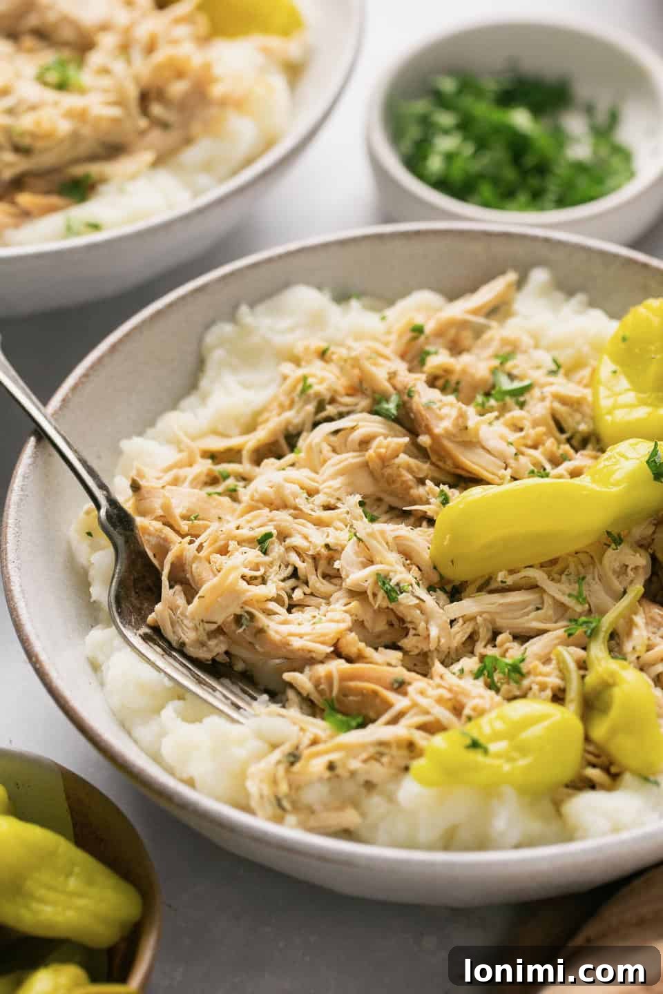 A white bowl filled with creamy mashed potatoes, topped with tender, shredded Mississippi chicken and vibrant pepperoncini peppers.