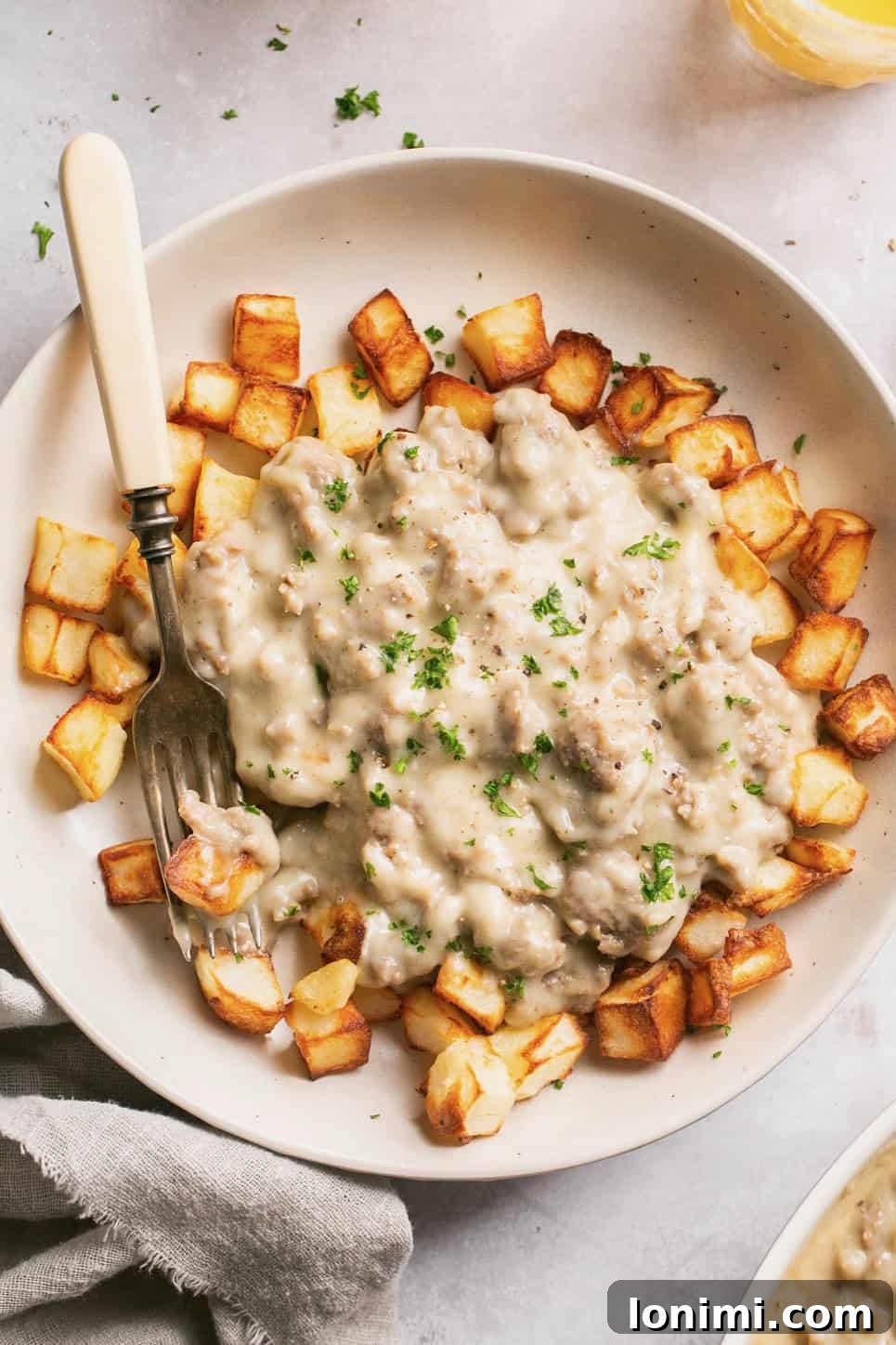 Effortless Keto Sausage Gravy 2 A close-up of creamy keto sausage gravy generously poured over crispy air-fried potatoes, with a fork resting beside the bowl, highlighting the delicious texture and rich flavor.