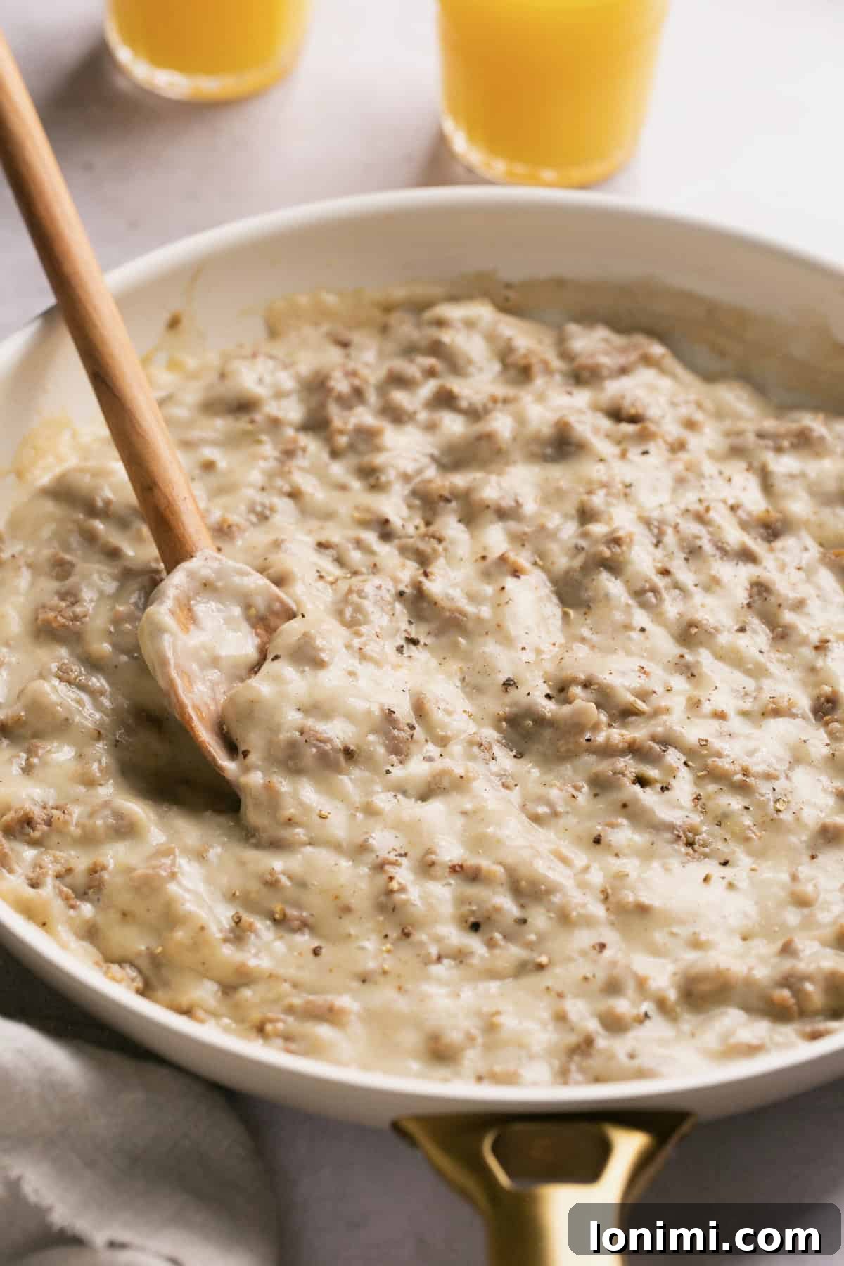 Effortless Keto Sausage Gravy 7 A wooden spoon gently stirring the creamy, golden-brown keto sausage gravy in a pristine white skillet, showcasing its smooth texture.