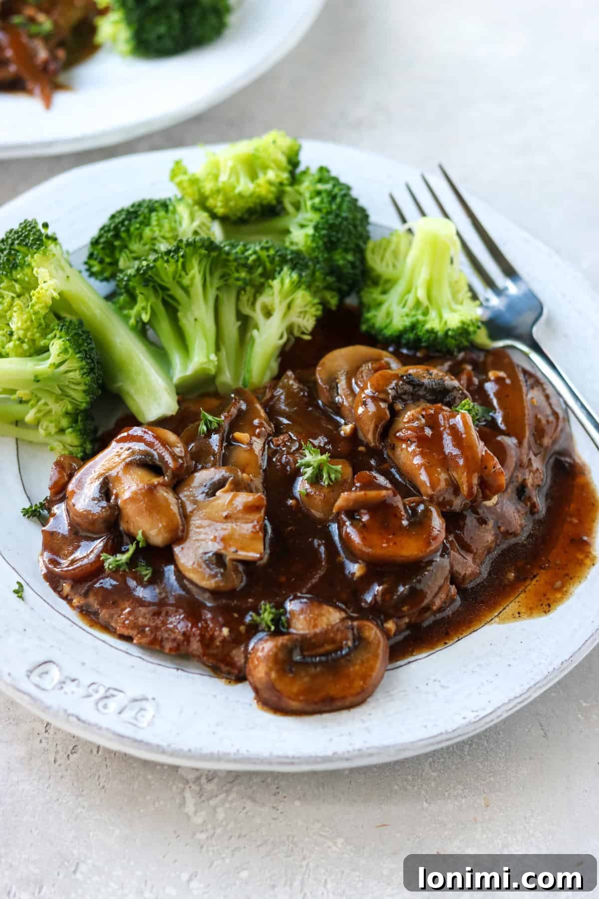 Effortless Tender Cube Steak 7 A serving of cube steak with gravy on a plate, adorned with sautéed mushrooms and accompanied by a side of steamed broccoli.