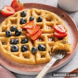 a gluten free waffle on a pink plate topped with blueberries, strawberries and maple syrup with a fork taking a bite out