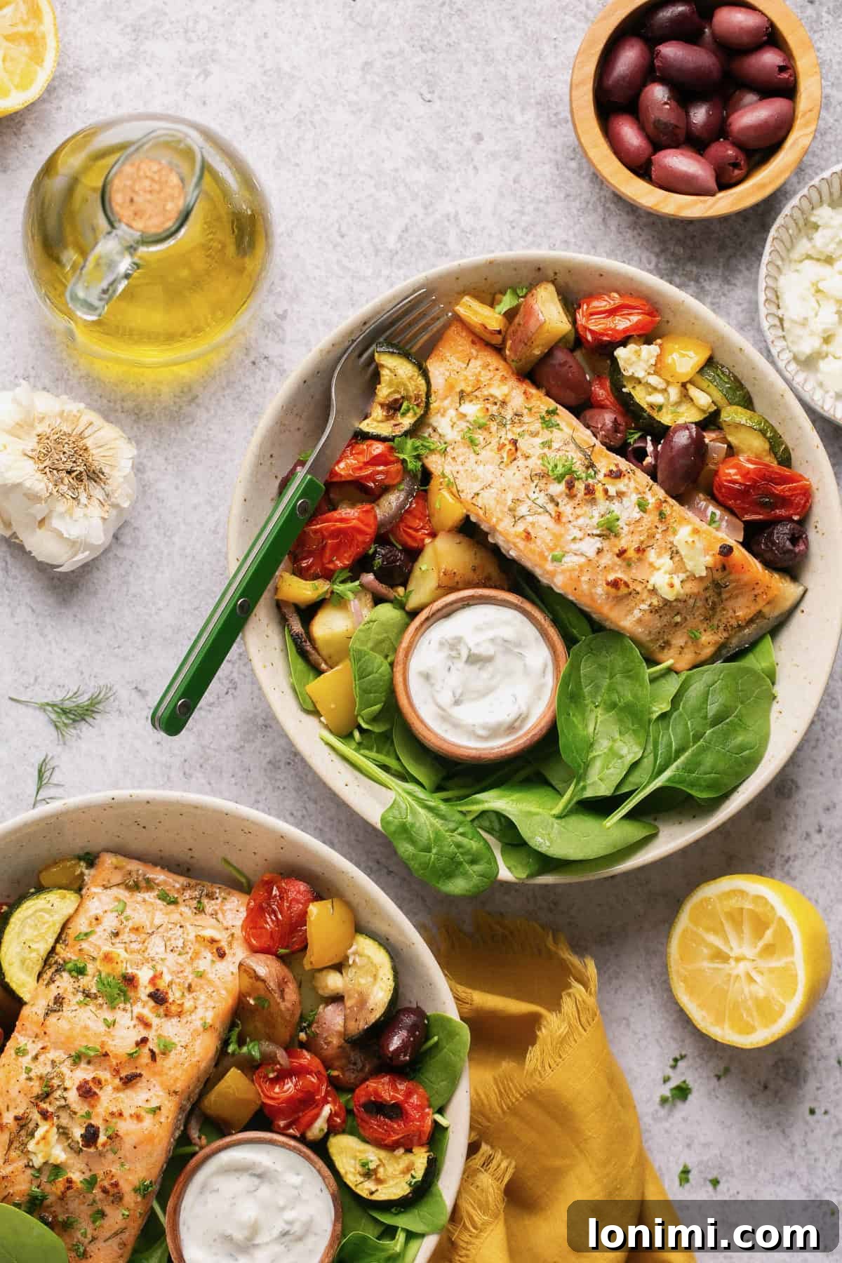 An overhead shot of two bowls of Greek salmon with veggies and spinach, accompanied by two small bowls of tzatziki sauce.