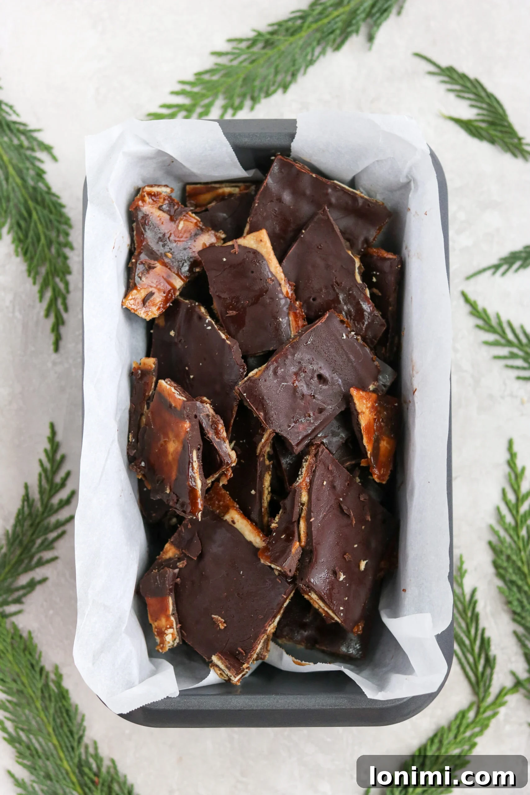 A festive loaf pan filled with a layer of cooled Paleo Christmas Crack, garnished with pine leaves and cinnamon sticks, ready for slicing.