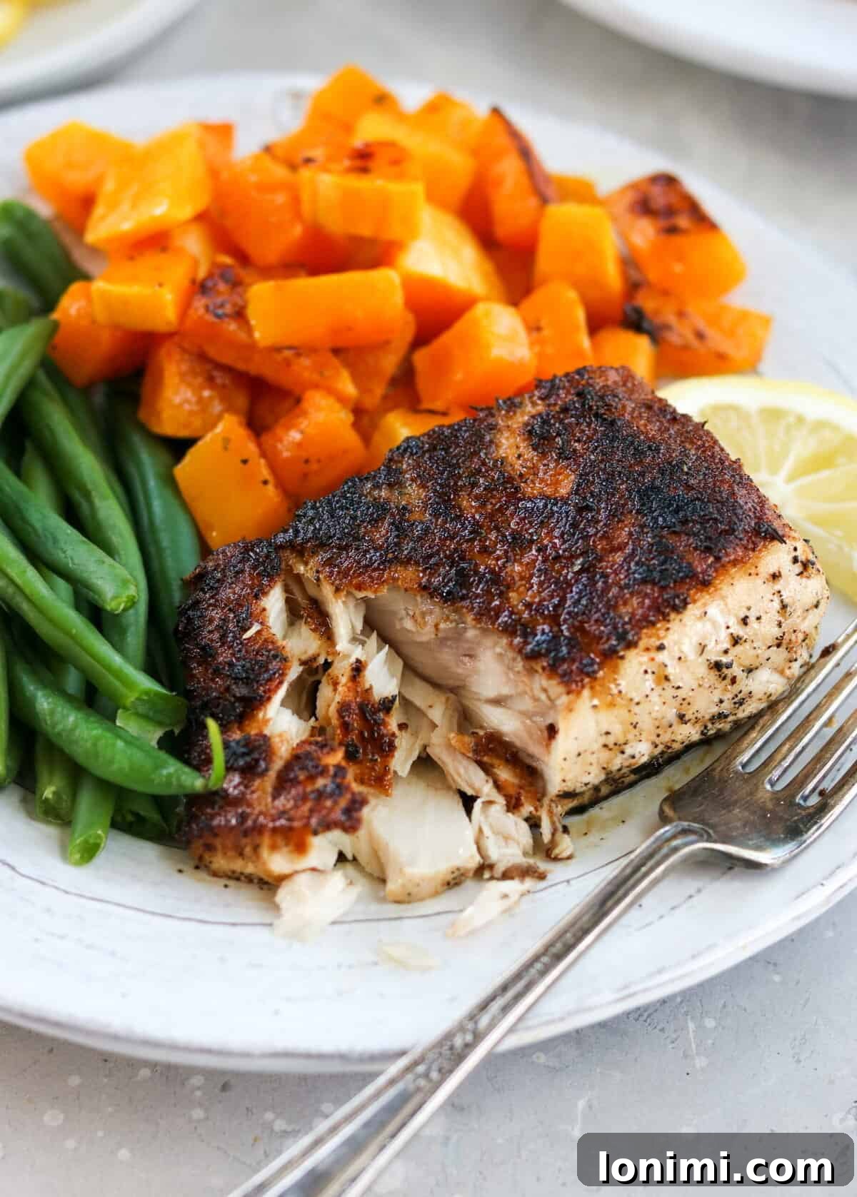 A perfectly cooked blackened mahi mahi fillet on a plate, garnished with fresh herbs and a wedge of lemon. The fish is flaking apart beautifully, served alongside roasted butternut squash and vibrant green beans, with a fork poised to enjoy.