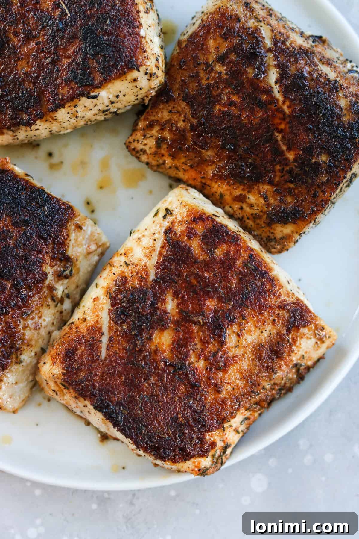 Four perfectly blackened Mahi Mahi fillets are arranged on a pristine white plate, showcasing their dark, crispy crust and tender interior, ready to be served.