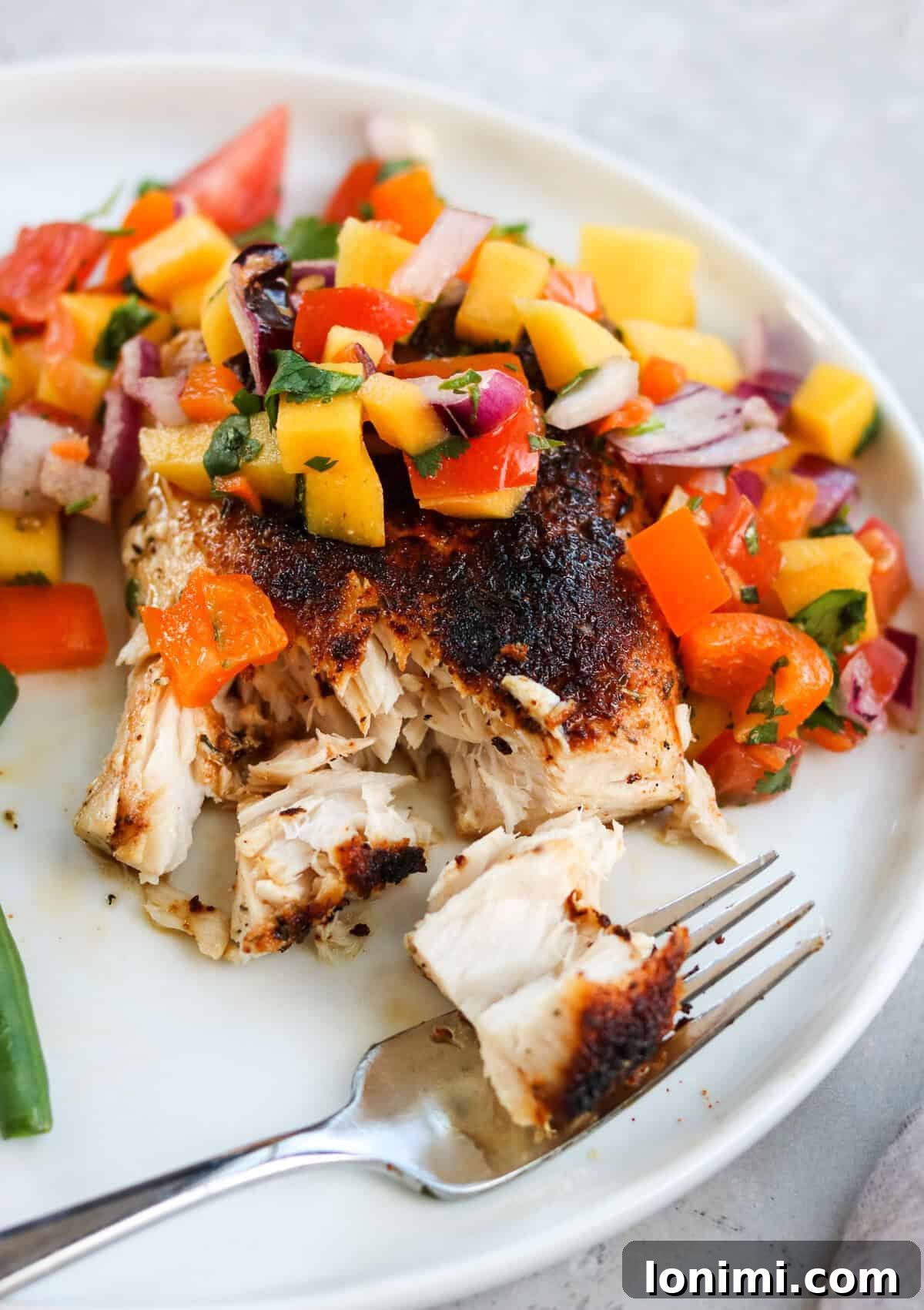 A perfectly cooked blackened fish fillet beautifully topped with a vibrant, colorful mango salsa, showcasing a delightful contrast of textures and flavors. The golden-brown fish is flaky and moist, complementing the fresh, chunky salsa.