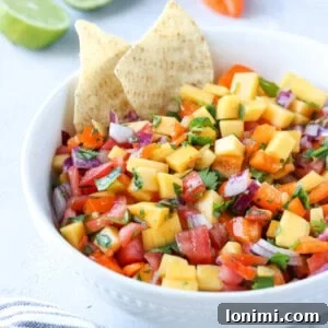 Irresistible Mango Habanero Salsa 6 A beautifully composed image of mango habanero salsa in a white bowl, with two tortilla chips ready for dipping. Fresh limes and a whole habanero pepper are artfully arranged in the background, hinting at the vibrant flavors within.