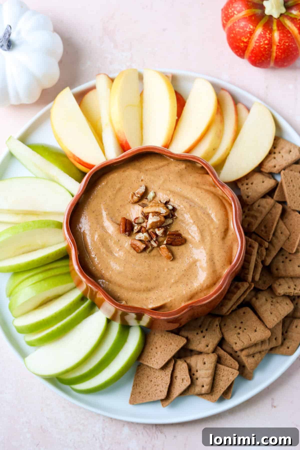 A charming pumpkin-shaped dish filled with creamy pumpkin dip, artfully arranged on a white plate with an assortment of fresh apple slices and gluten-free graham crackers for dipping.