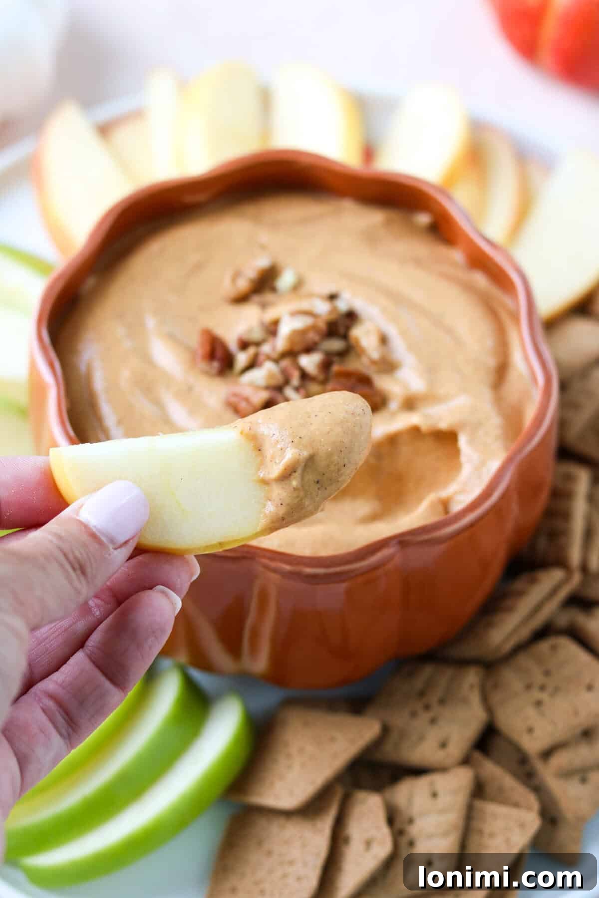 A hand reaches into a pumpkin pie dip with a fresh apple slice, illustrating the delicious and inviting nature of the healthy fall dessert or appetizer.