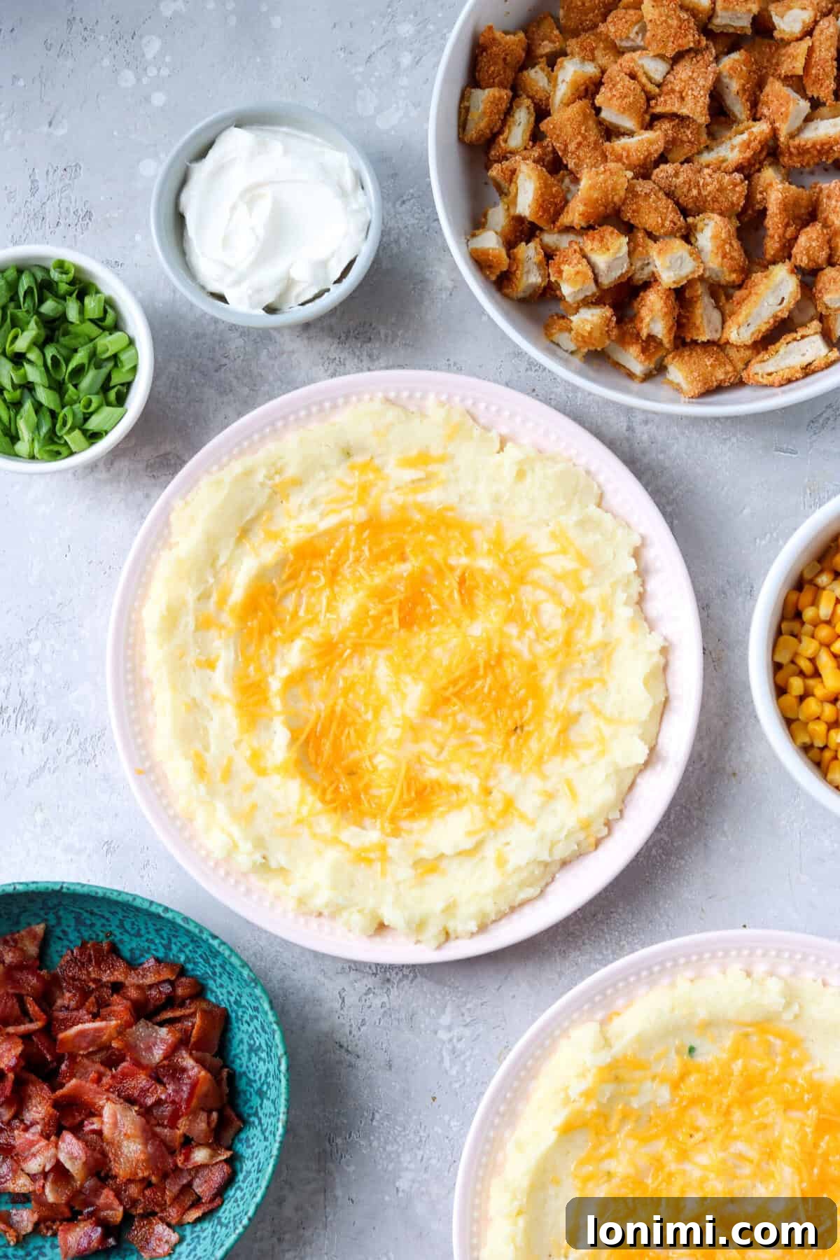 Ultimate Mashed Potato Bowls 6 a bowl of mashed potatoes topped with cheese surrounded by bowls of chicken, sour cream, green onions, bacon, and corn