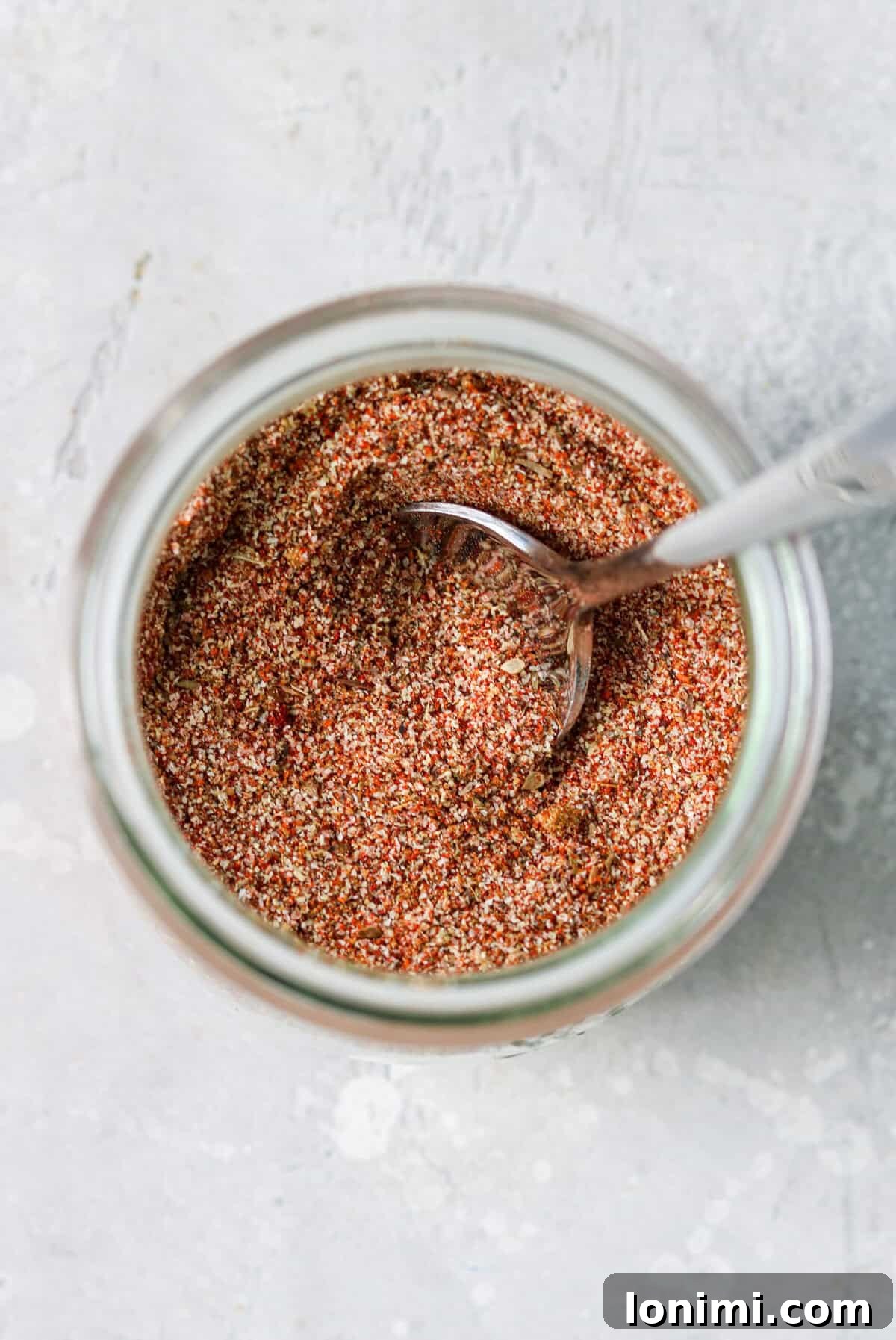 A clear glass jar filled with homemade chicken taco seasoning, with a silver spoon resting inside. The background suggests a rustic kitchen setting.