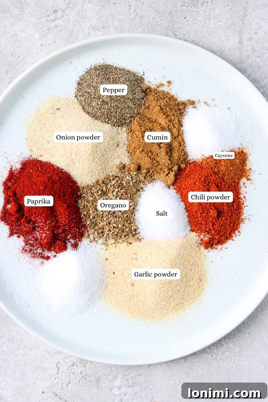 Individual mounds of various taco seasoning spices laid out on a white plate with small labels, including chili powder, cumin, and paprika.