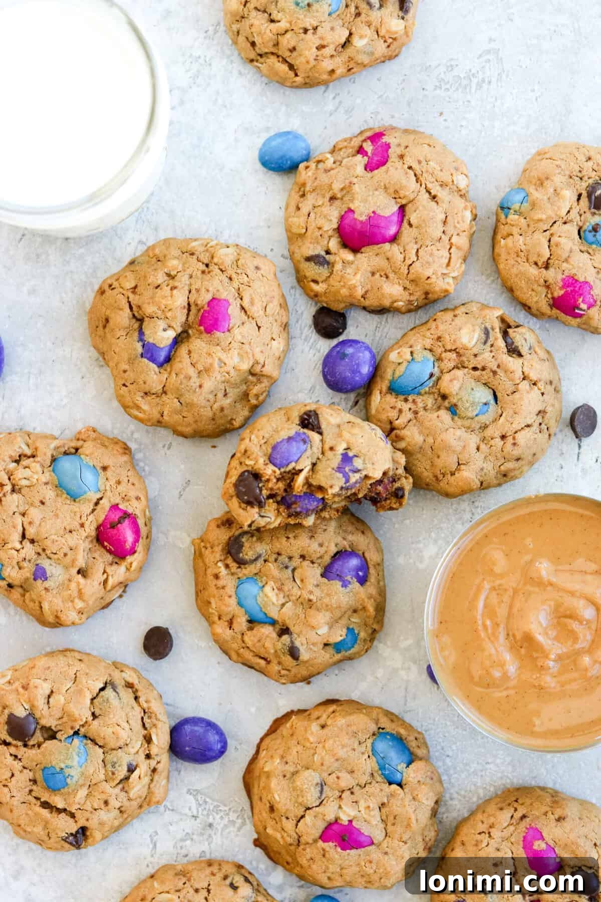 A perfectly stacked trio of gluten free monster cookies, brimming with colorful chocolate candies and dark chocolate chips, accompanied by a glass of refreshing milk and a bowl of creamy peanut butter. The cookies showcase their soft, chewy texture and vibrant inclusions, inviting a closer look.