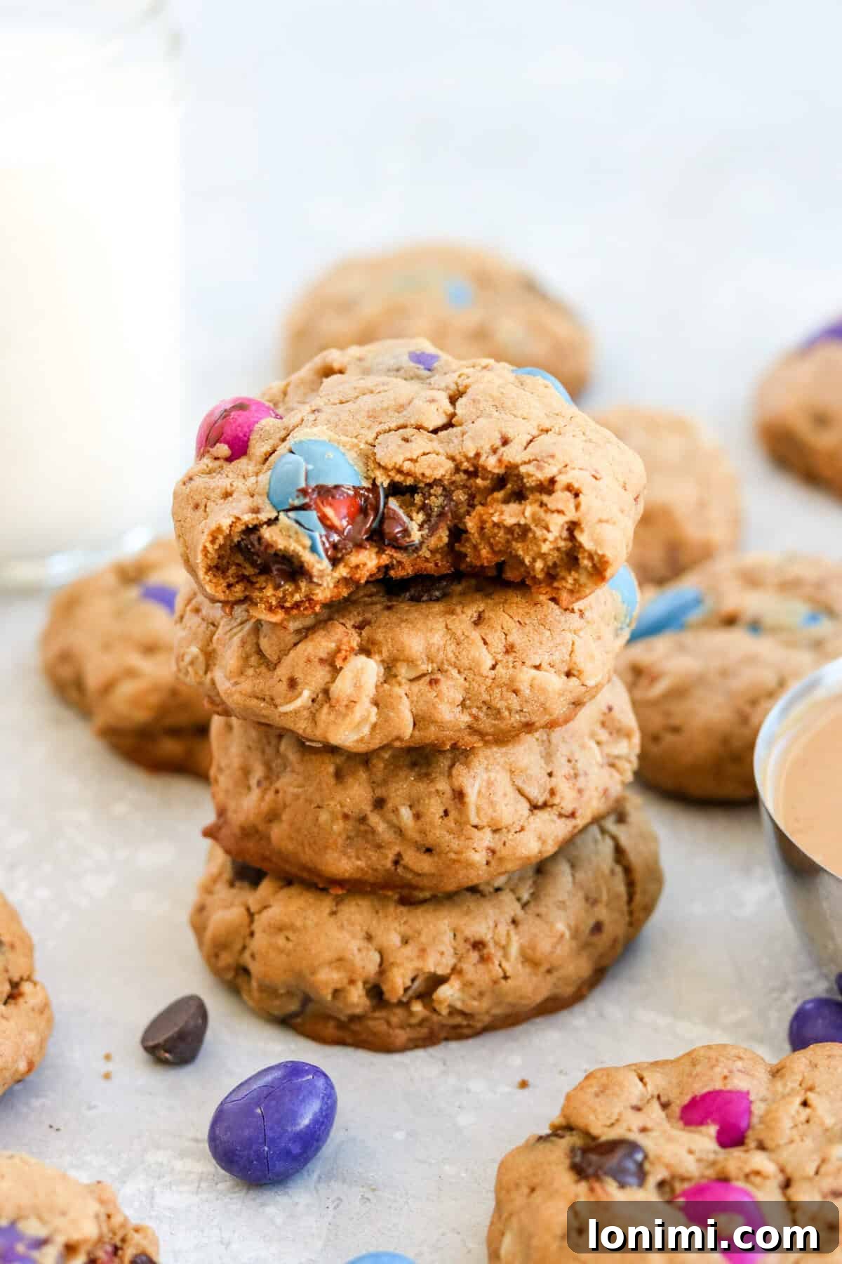 A tantalizing stack of four gluten free monster cookies, surrounded by scattered M&M's and other cookies, emphasizing their deliciousness and abundance. The cookies are rich in color and texture, showcasing their generous fillings.